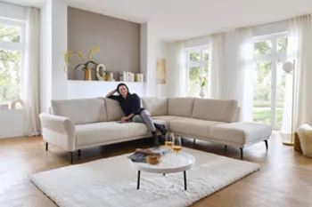 Auf dem Bild ist ein Wohnzimmer zu sehen. Eine helle, moderne Couch mit schwarzen Füßen. Es handelt sich hier um eine L Couch. Vor der Couch steht ein Couchtisch und darunter liegt ein schöner flauschiger, heller Teppich.